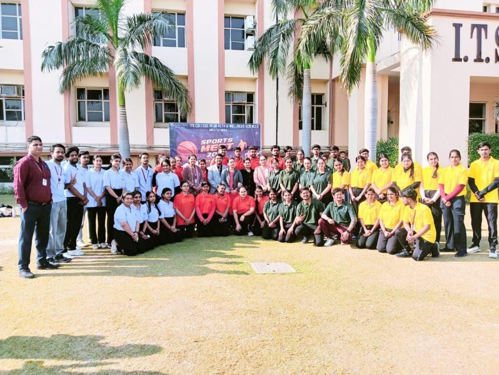 Students and faculty members of I.T.S College of Health & Wellness Sciences, Greater Noida, pose together on the college lawn during the Sports Meet 2024, wearing color-coded uniforms representing different teams, with a banner in the background and the college building visible.
